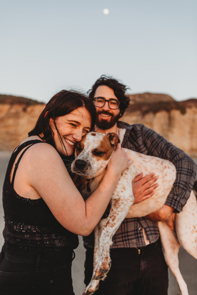 couple at beach with dog kissing dog