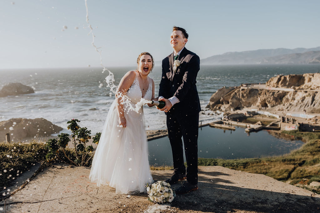 couple popping champagne on ocean cliff side during elopement